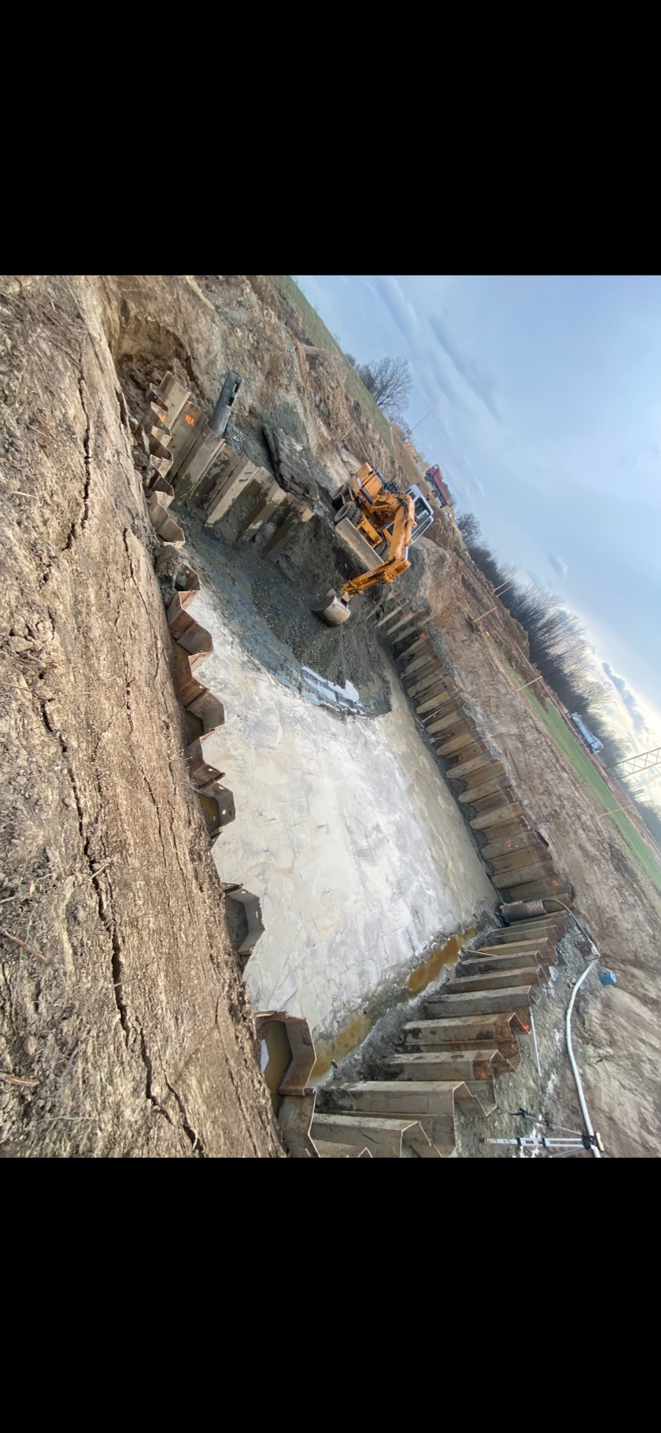 Żółta koparka w wykopie zabezpieczonym stalowymi szalunkami, częściowo wypełnionym wodą, widok z góry na plac budowy.