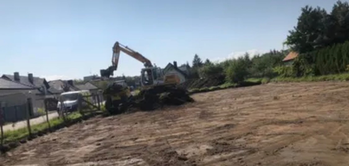 Żółta koparka podczas prac ziemnych na działce budowlanej, w tle zabudowa jednorodzinna i ogrodzenie z siatki.
