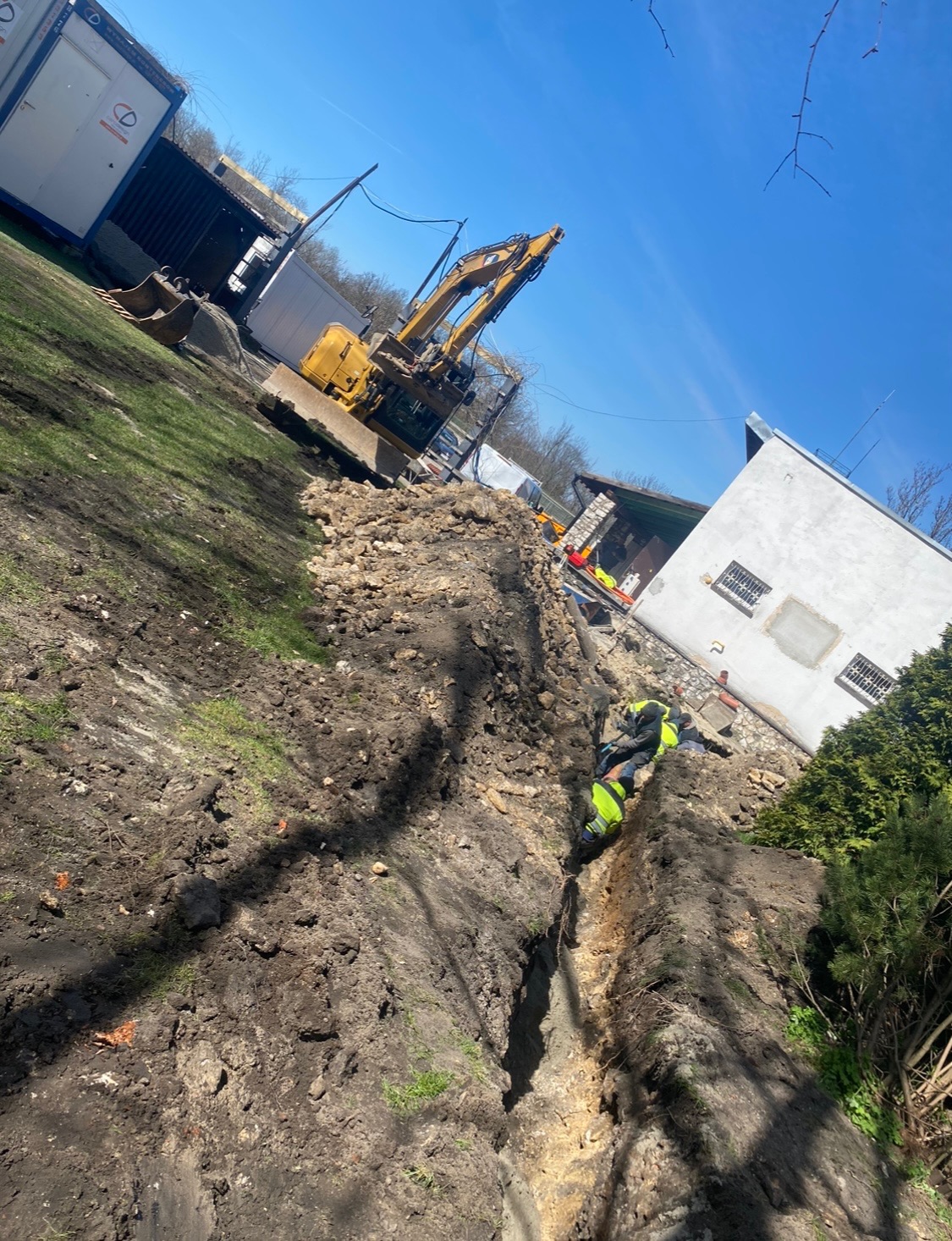 Żółta koparka na placu budowy, obok wykopany wąski rów, w którym pracuje dwóch robotników w jaskrawych kamizelkach odblaskowych, w tle budynek i kontenery.
