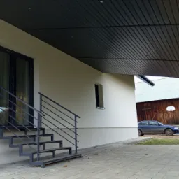 Nowoczesny dom z jasną elewacją, czarnymi ramami okiennymi i schodami z metalową barierką; w tle drewniana stodoła z koszem do koszykówki.