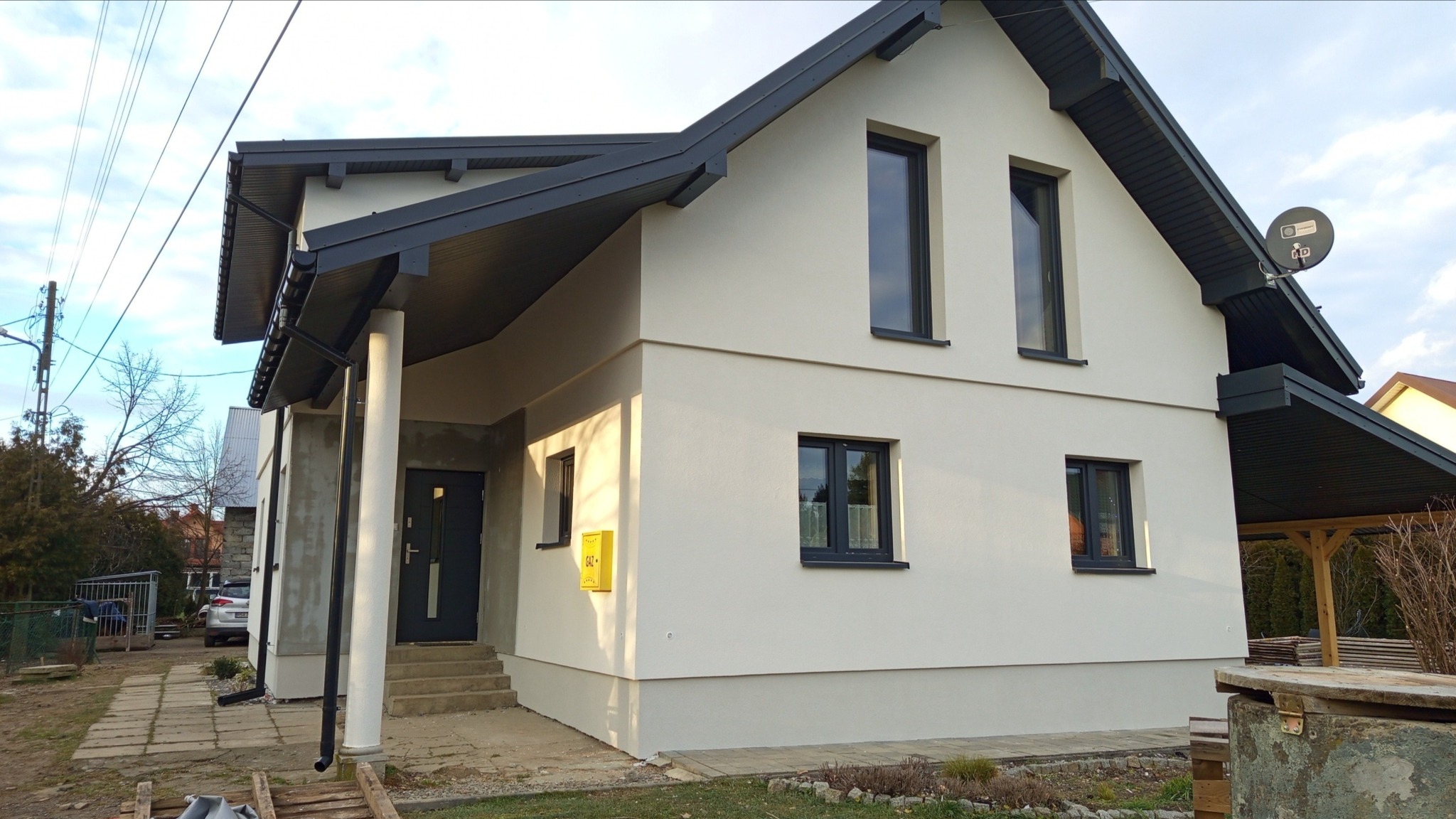 Dom jednorodzinny z jasną elewacją, ciemnymi oknami i dachem, widoczny z zewnątrz, z żółtą skrzynką gazową na ścianie i anteną satelitarną na dachu.