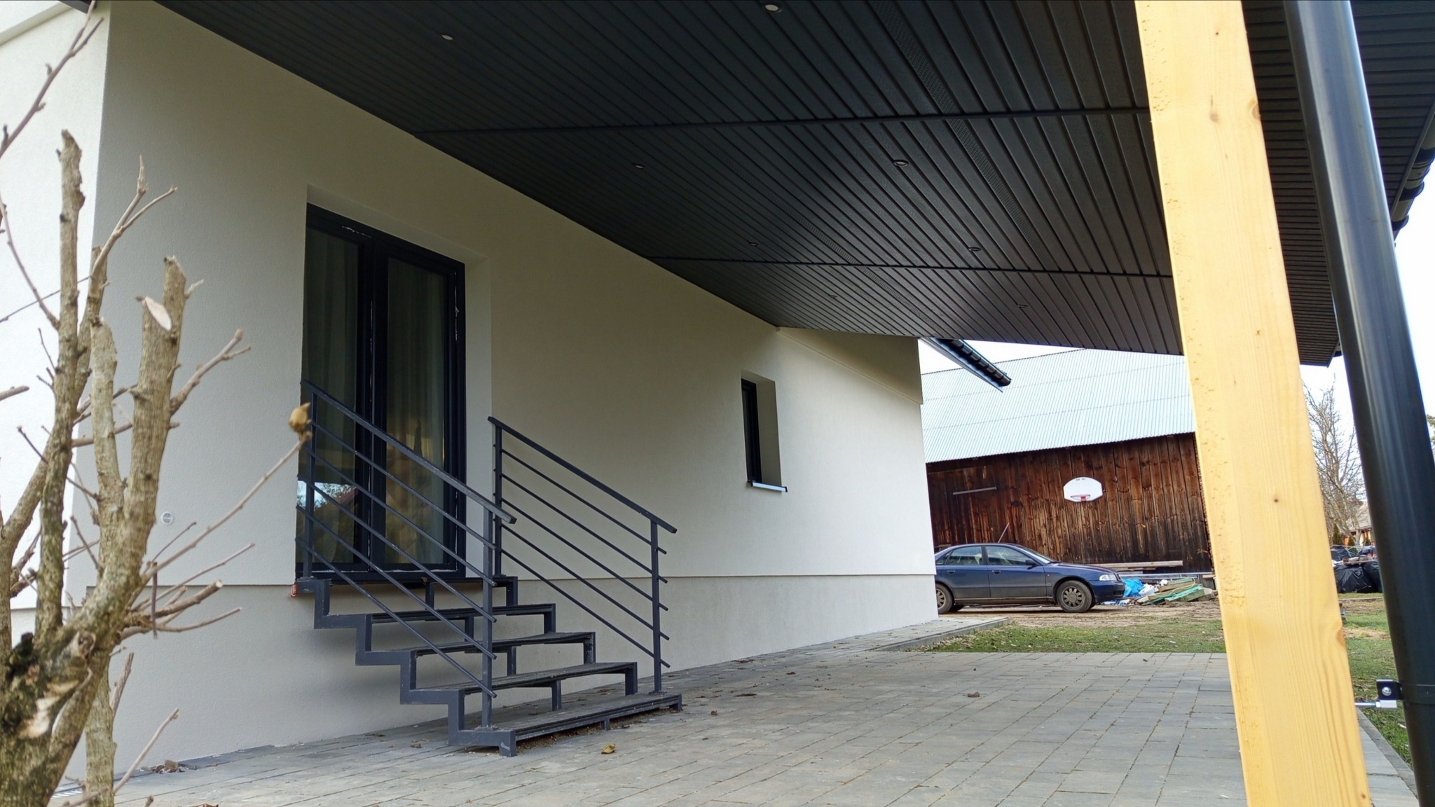 Nowoczesny dom z jasną elewacją, czarnymi ramami okiennymi i schodami z metalową barierką; w tle drewniana stodoła z koszem do koszykówki.