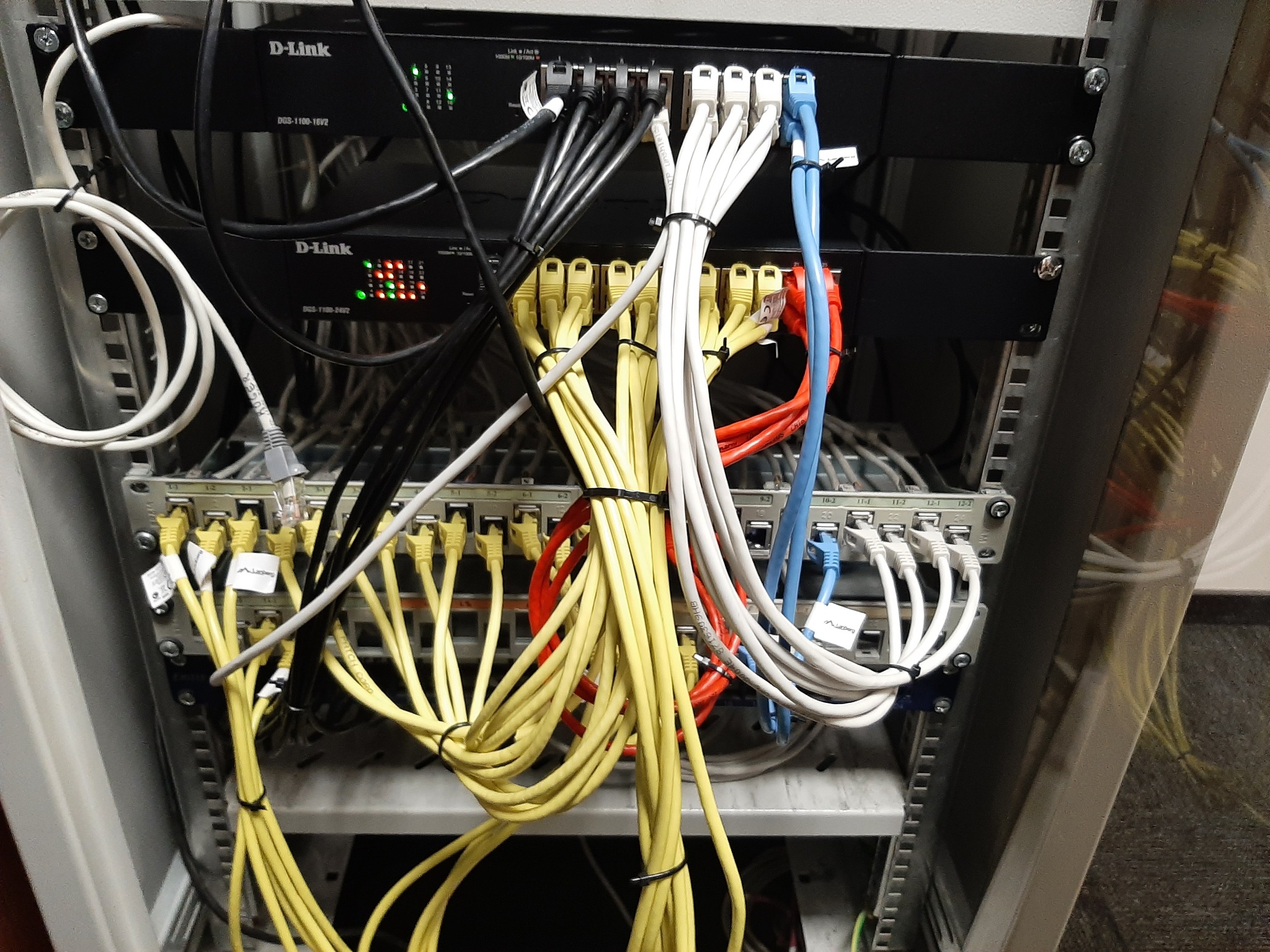 Szafa serwerowa z zainstalowanymi dwoma switchami D-Link, okablowanie sieciowe w kolorach żółtym, białym, czarnym, niebieskim i czerwonym, kable spięte opaskami zaciskowymi, widoczne diody LED...