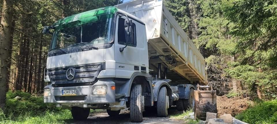 Ciężarówka Mercedes-Benz 4141 z podniesioną skrzynią ładunkową, zaparkowana na leśnej drodze, prawdopodobnie podczas wywozu ziemi lub gruzu. Widoczne drzewa w tle.