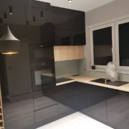 Nowoczesna, czarna kuchnia z połyskiem w Rogoźnie, z drewnianym blatem i oświetleniem punktowym, widoczny piekarnik i mikrofalówka w zabudowie oraz regał na wino.