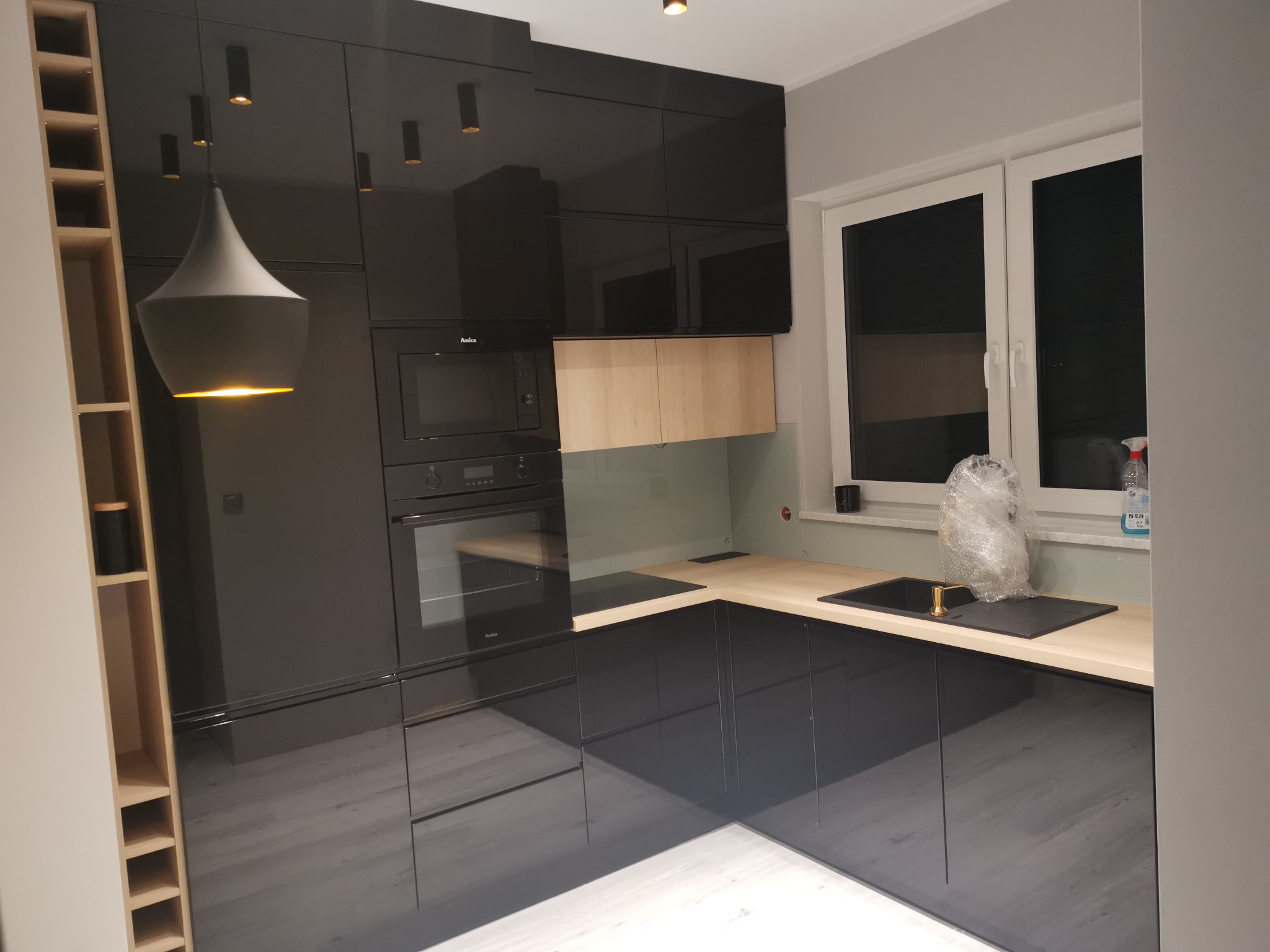 Nowoczesna, czarna kuchnia z połyskiem w Rogoźnie, z drewnianym blatem i oświetleniem punktowym, widoczny piekarnik i mikrofalówka w zabudowie oraz regał na wino.