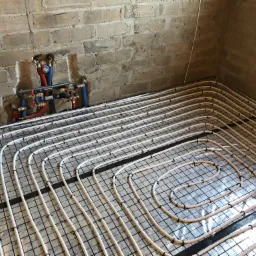 Rozłożona instalacja ogrzewania podłogowego na siatce, widoczne rury i rozdzielacz na tle surowej ściany z cegły.