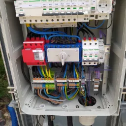 Otwarta skrzynka elektryczna z widocznymi bezpiecznikami, przewodami o różnych kolorach i elementami ochronnymi, zamontowana na zewnątrz budynku.