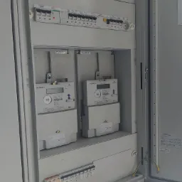 Otwarta szara szafa elektryczna z dwoma licznikami energii Norax 3, zabezpieczeniami nadprądowymi EATON i zielono-żółtym przewodem uziemiającym. Widoczne naklejki informacyjne i tabliczki znamionowe.