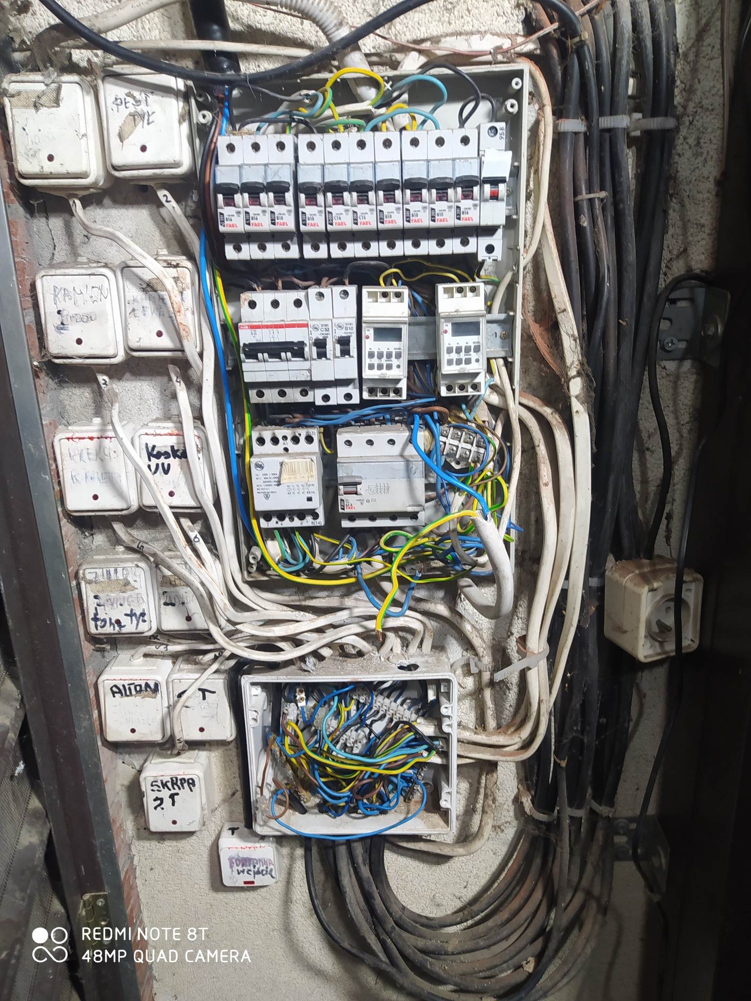 Stara, zaniedbana skrzynka bezpiecznikowa z licznymi przewodami, wyłącznikami i przełącznikami, niektóre z napisami odręcznymi na ścianie, widoczne ślady użytkowania i zabrudzenia.