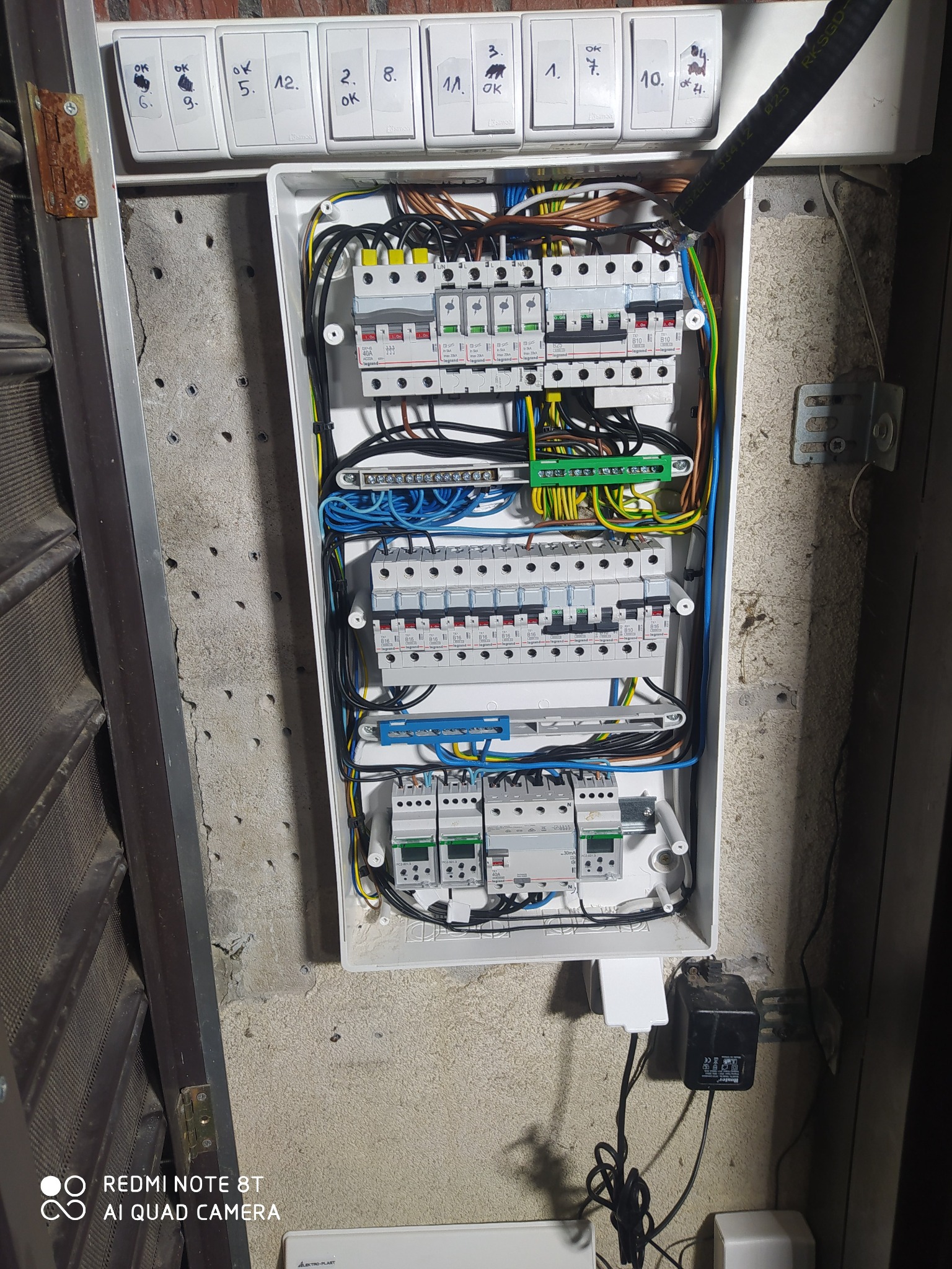 Otwarta skrzynka rozdzielcza z widocznymi bezpiecznikami, przewodami i listwami zaciskowymi, zamontowana na surowej ścianie z widocznymi śladami po wierceniu, z podpisami nad wyłącznikami...