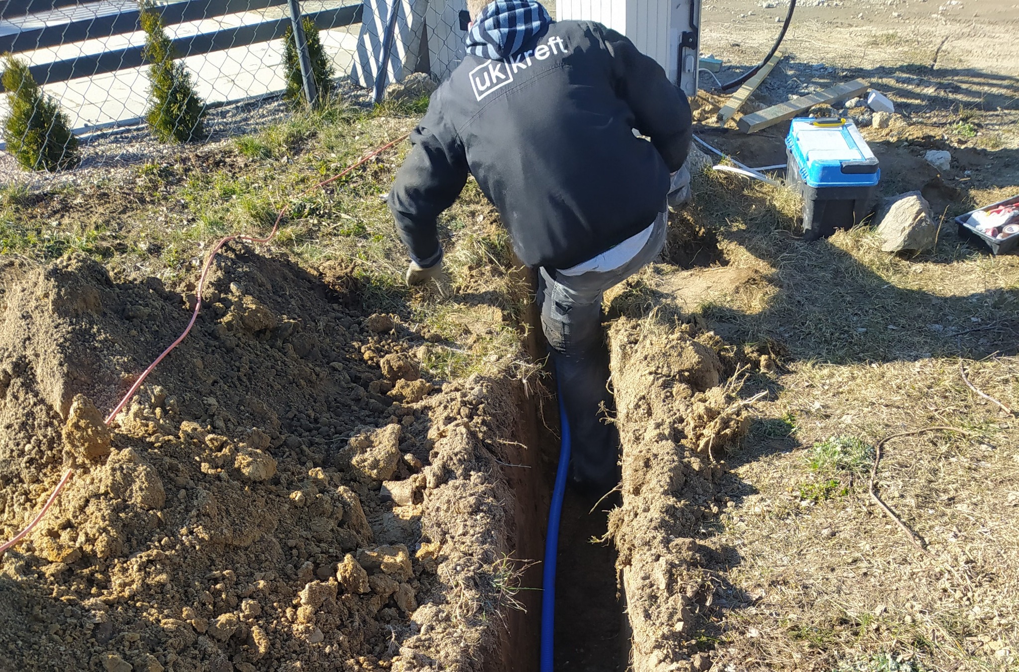 Pracownik w kurtce z logo firmy umieszcza niebieski przewód w wykopanym rowie w ziemi, obok leży skrzynka z narzędziami.