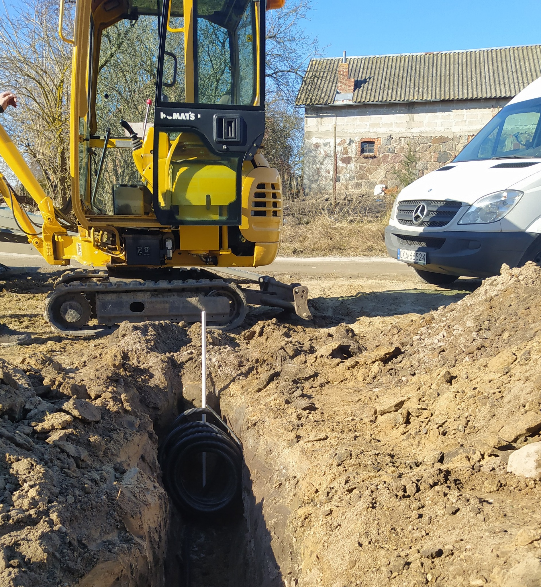 Żółta mini koparka obok wykopu z czarnym karbowanym przewodem w ziemi, na tle biały van i budynek z cegły.