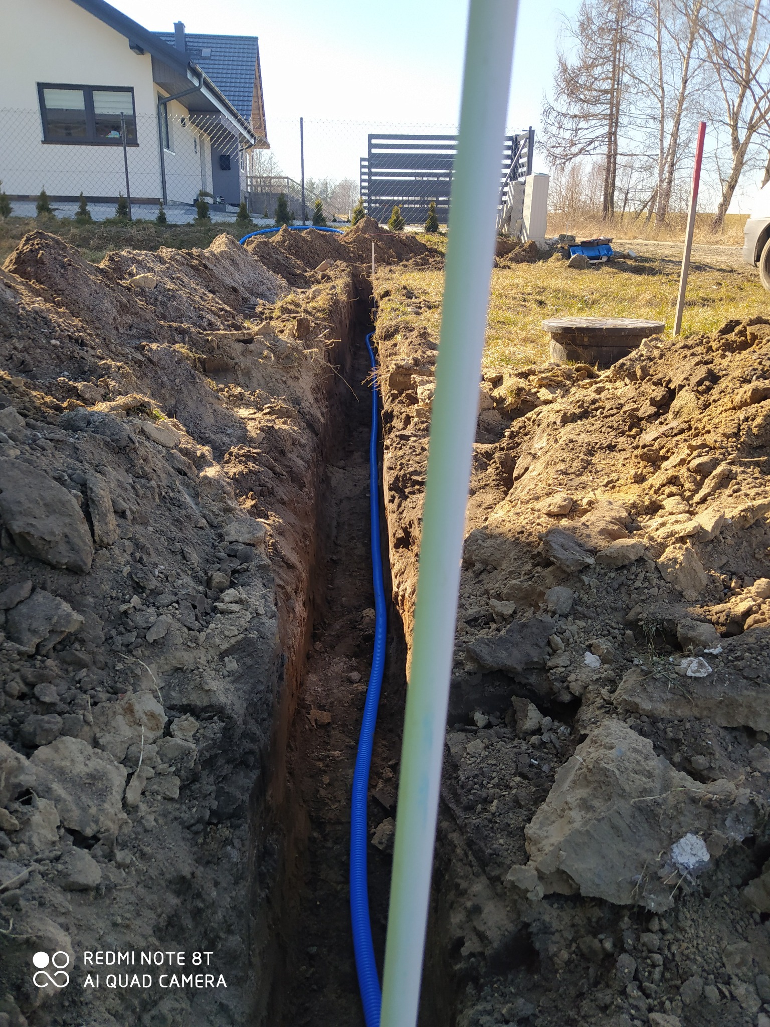 Wąski wykop w ziemi z ułożoną niebieską rurą karbowaną, prawdopodobnie do kabli elektrycznych, widok z góry z przesłonięciem przez biały element instalacji, w tle dom jednorodzinny i drzewa.