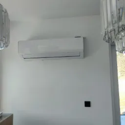 Biały klimatyzator ścienny zamontowany w nowoczesnym wnętrzu, widoczne kryształowe lampy wiszące i czarny panel sterowania na ścianie.