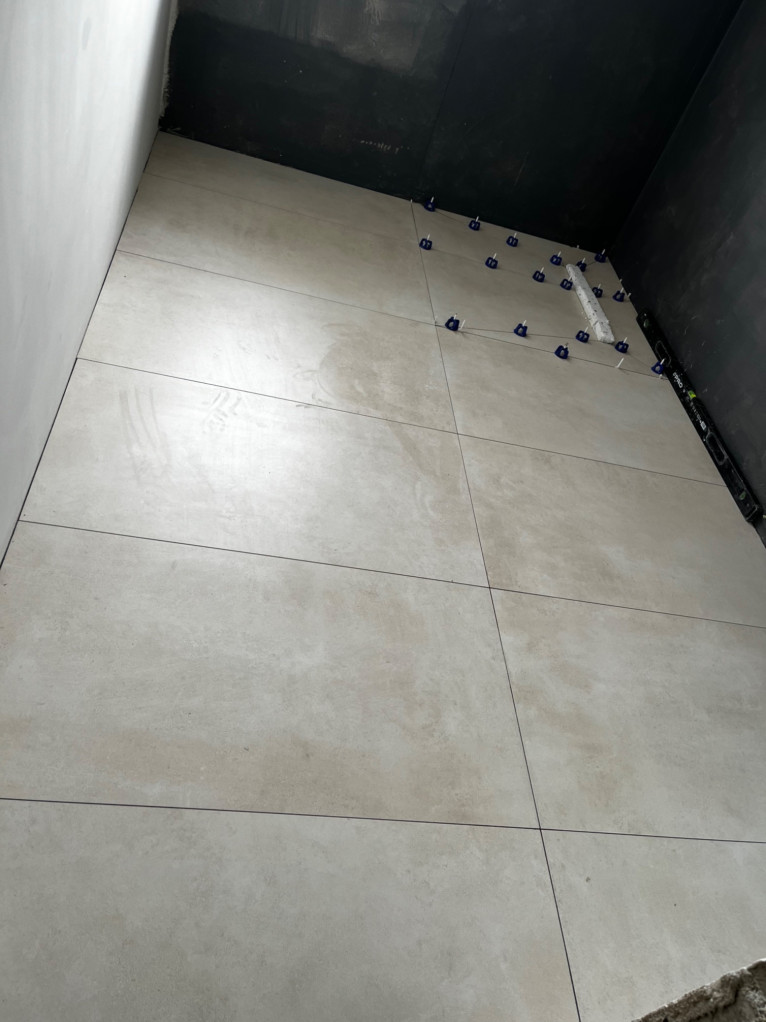 Układanie jasnych, wielkoformatowych płytek na podłodze z użyciem klipsów poziomujących, widoczny fragment rury i poziomica przy ścianie pomalowanej na czarno.