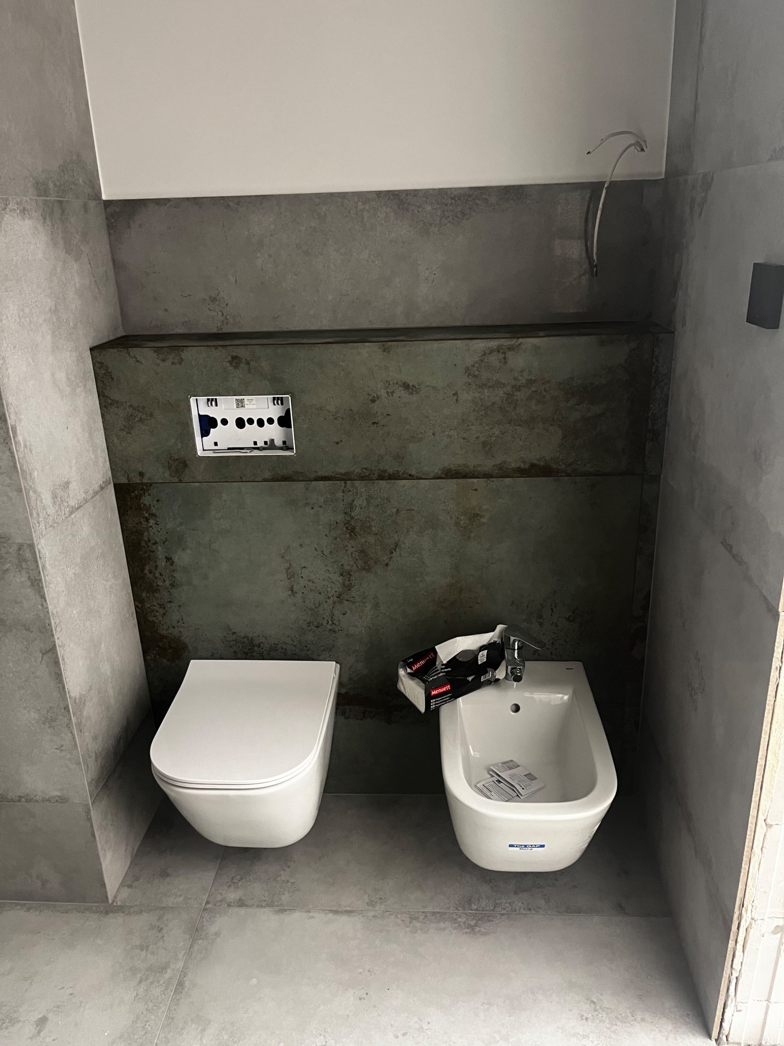 Nowoczesna łazienka z szarymi płytkami imitującymi beton, wisząca toaleta i bidet, widoczne elementy instalacji oraz opakowanie armatury.