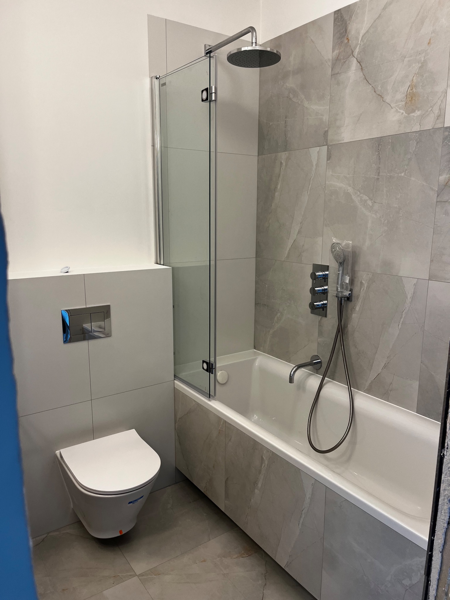 Nowoczesna łazienka z wanną obudowaną płytkami imitującymi marmur, szklanym parawanem nawannowym, podwieszaną toaletą i armaturą w kolorze chromu, jeszcze zafoliowana słuchawka prysznicowa.