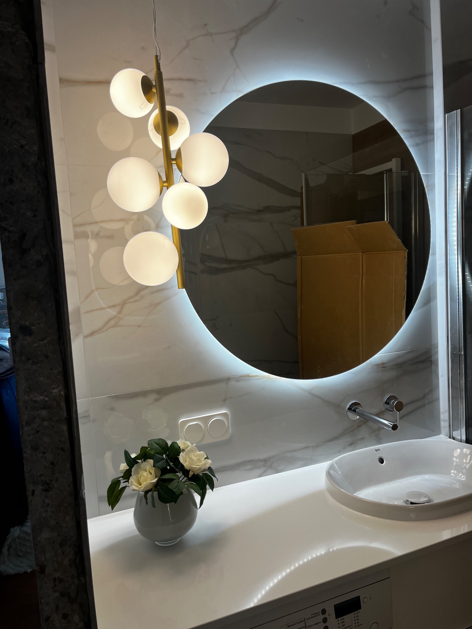 Nowoczesna łazienka z okrągłym lustrem podświetlanym LED, marmurową ścianą, designerską lampą kulową, umywalką nablatową i kwiatami w wazonie.