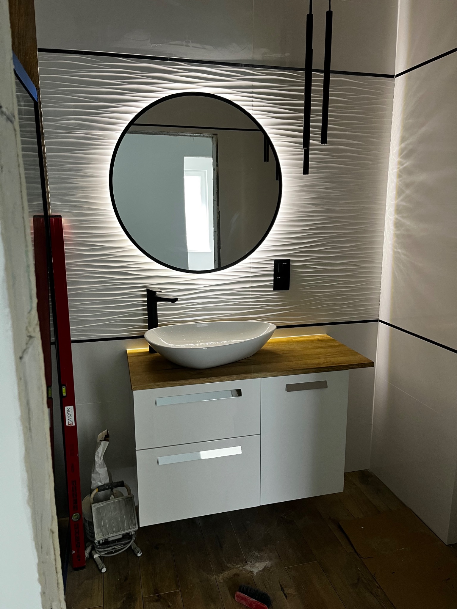 Nowoczesna łazienka w trakcie wykańczania: owalne umywalka na drewnianym blacie, podświetlane okrągłe lustro na ścianie z falistych płytek, widoczny poziomica i lampa halogenowa.