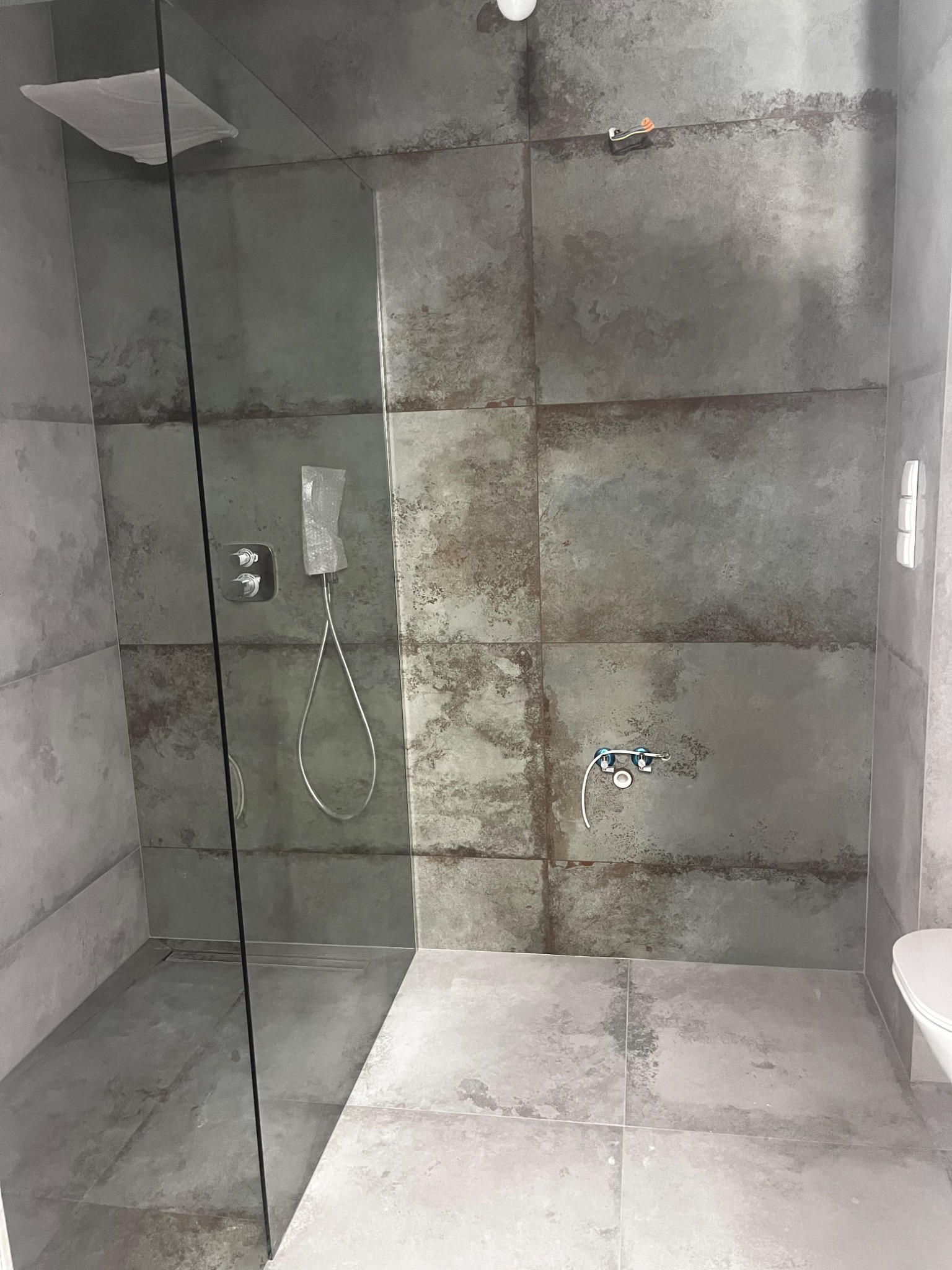 Kabina prysznicowa walk-in z szarymi płytkami imitującymi beton, widoczne przyłącza wodne i odpływ liniowy w podłodze.