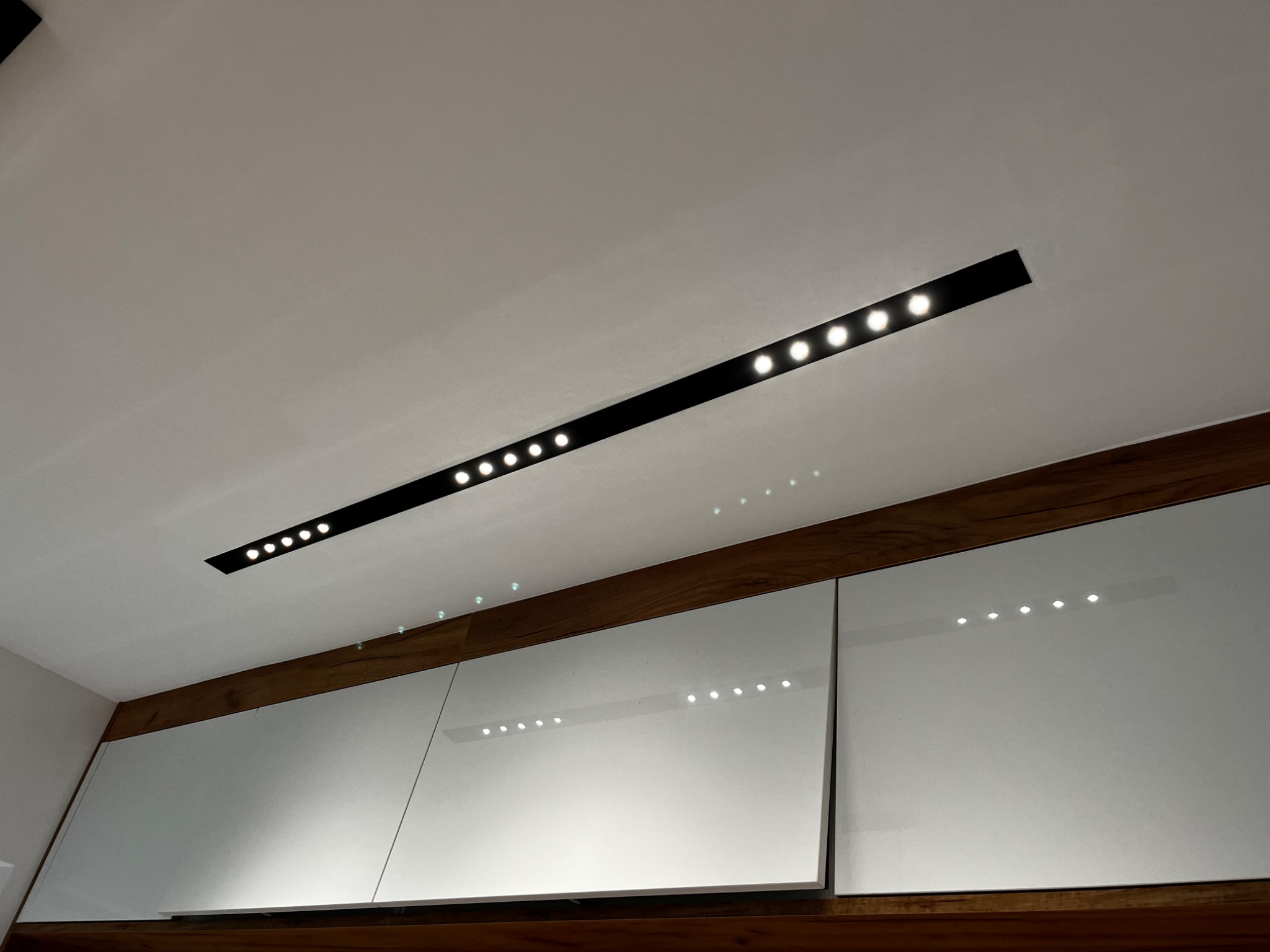 Nowoczesne oświetlenie sufitowe z wbudowanymi punktami świetlnymi, odbijające się w gładkich, białych frontach szafek kuchennych z drewnianymi akcentami.