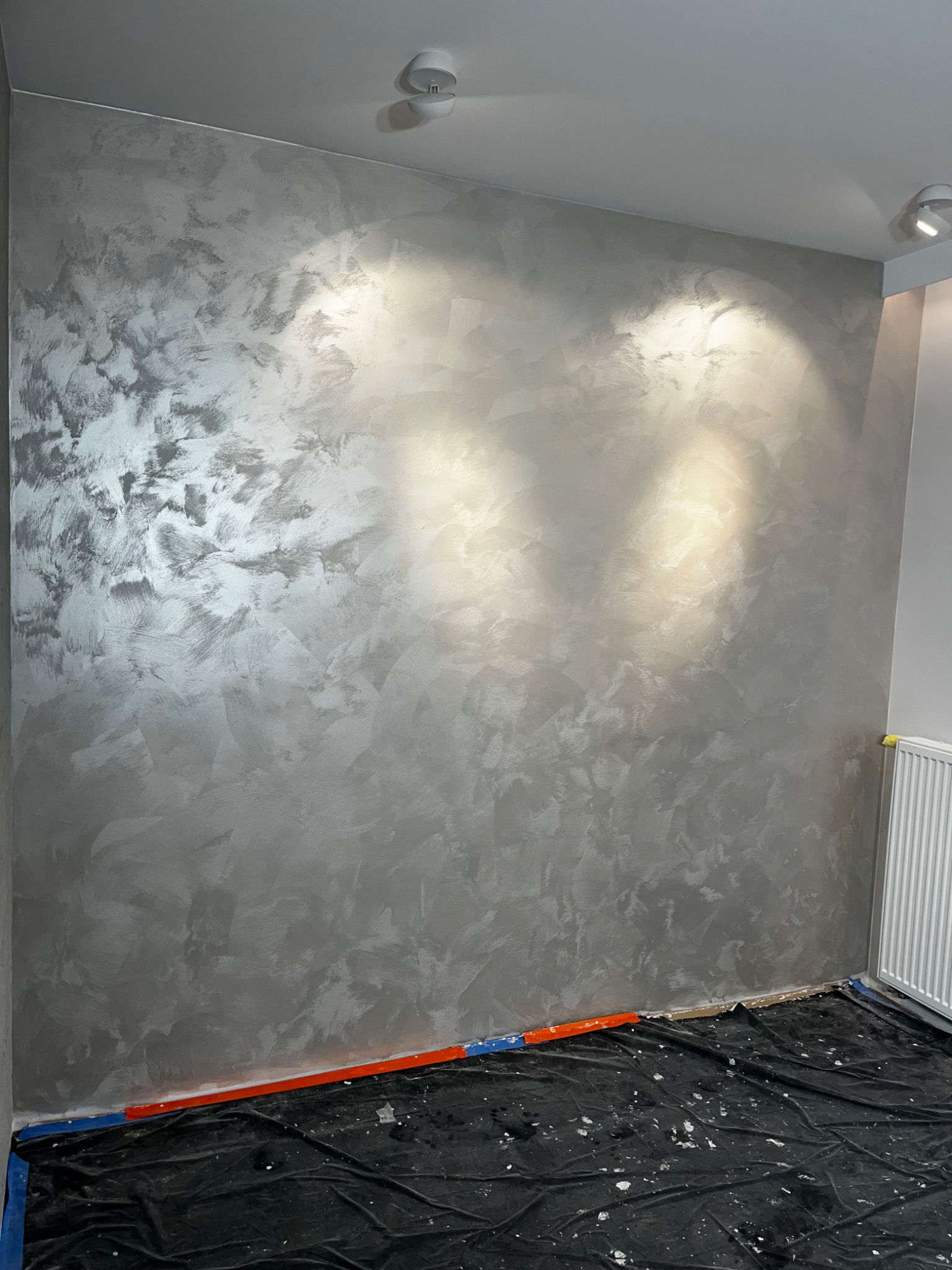 Ściana w pokoju pokryta dekoracyjną, srebrzysto-szarą gładzią w stylu weneckim, z widocznymi śladami pędzla i refleksami światła, zabezpieczona folią malarską na podłodze i grzejnikiem w rogu.