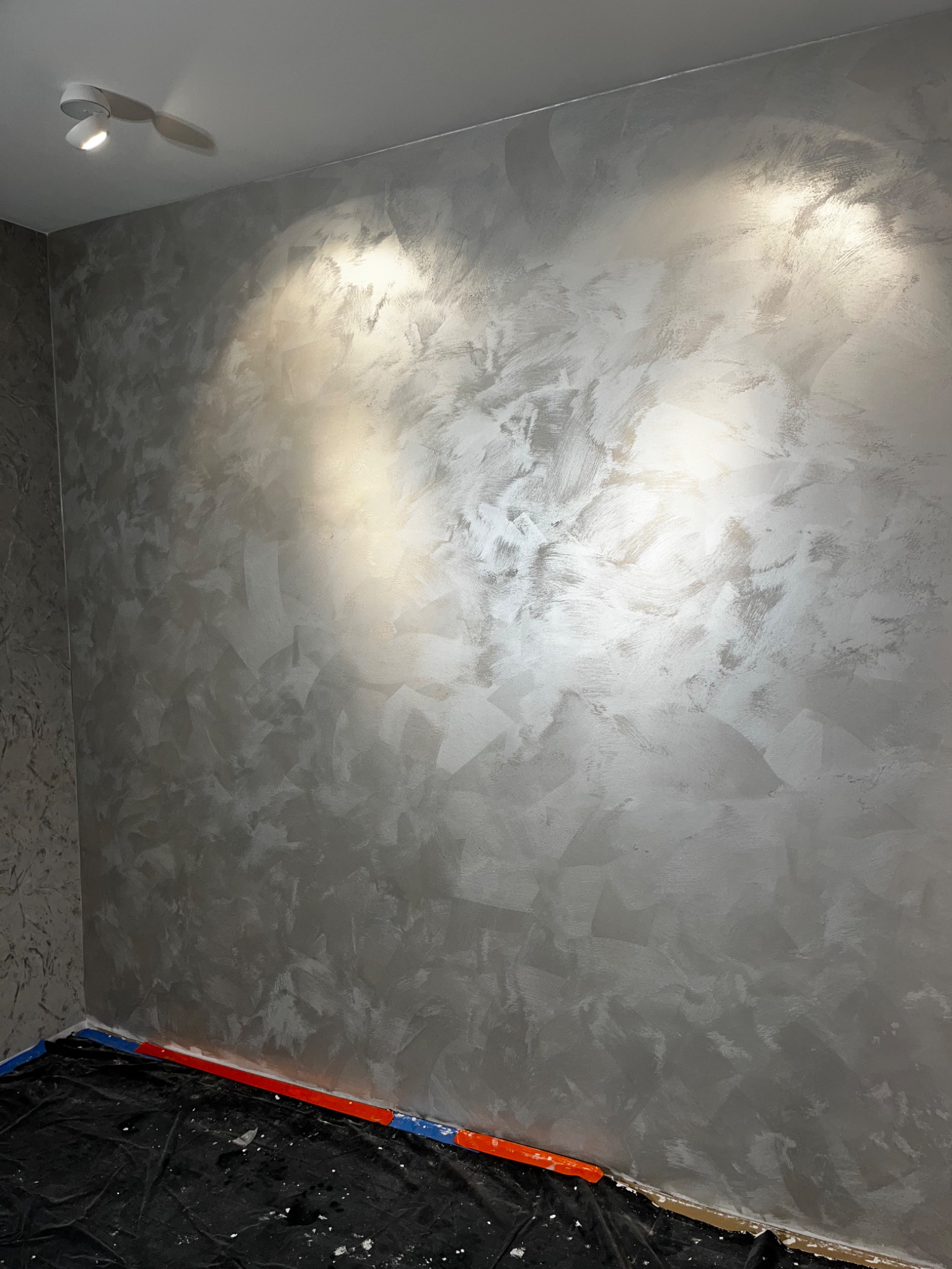 Ściana w pokoju po nałożeniu dekoracyjnej, srebrzystej gładzi, z widocznymi śladami pędzla i zabezpieczoną folią podłogą.
