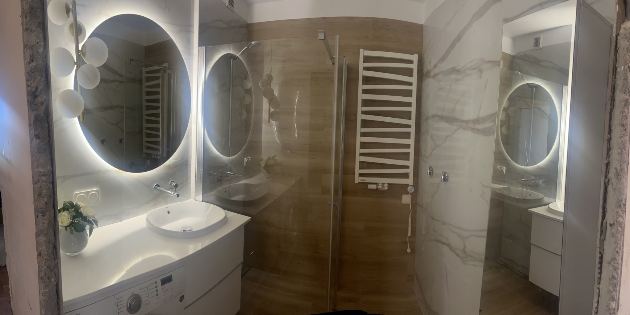 Nowoczesna łazienka z okrągłymi lustrami podświetlanymi LED, kabiną prysznicową ze szklanymi drzwiami i grzejnikiem drabinkowym, wykończona płytkami imitującymi marmur i drewno.
