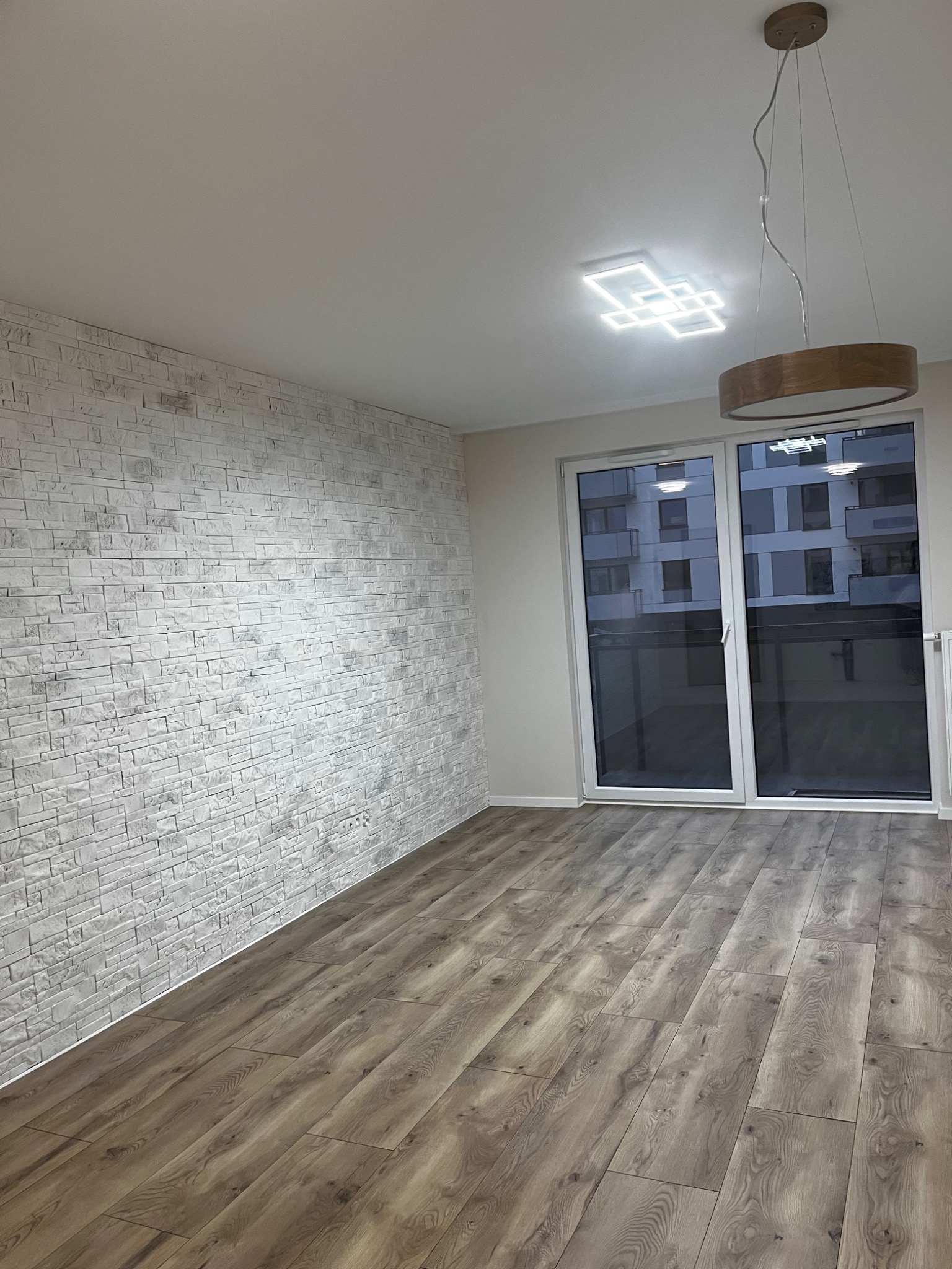 Pusty pokój po remoncie z panelami podłogowymi w odcieniach brązu, ścianą wykończoną białą cegłą dekoracyjną, dużym oknem balkonowym i nowoczesnym oświetleniem sufitowym.