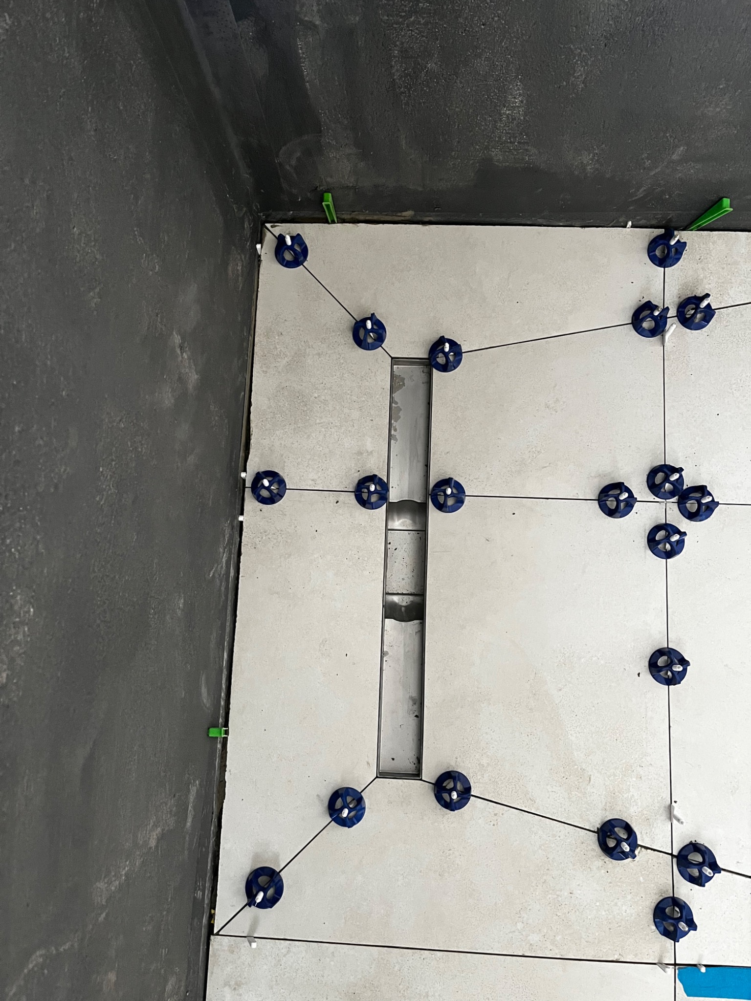 Ułożone płytki podłogowe w trakcie montażu z użyciem systemu poziomowania, widoczny odpływ liniowy i surowa ściana w tle.