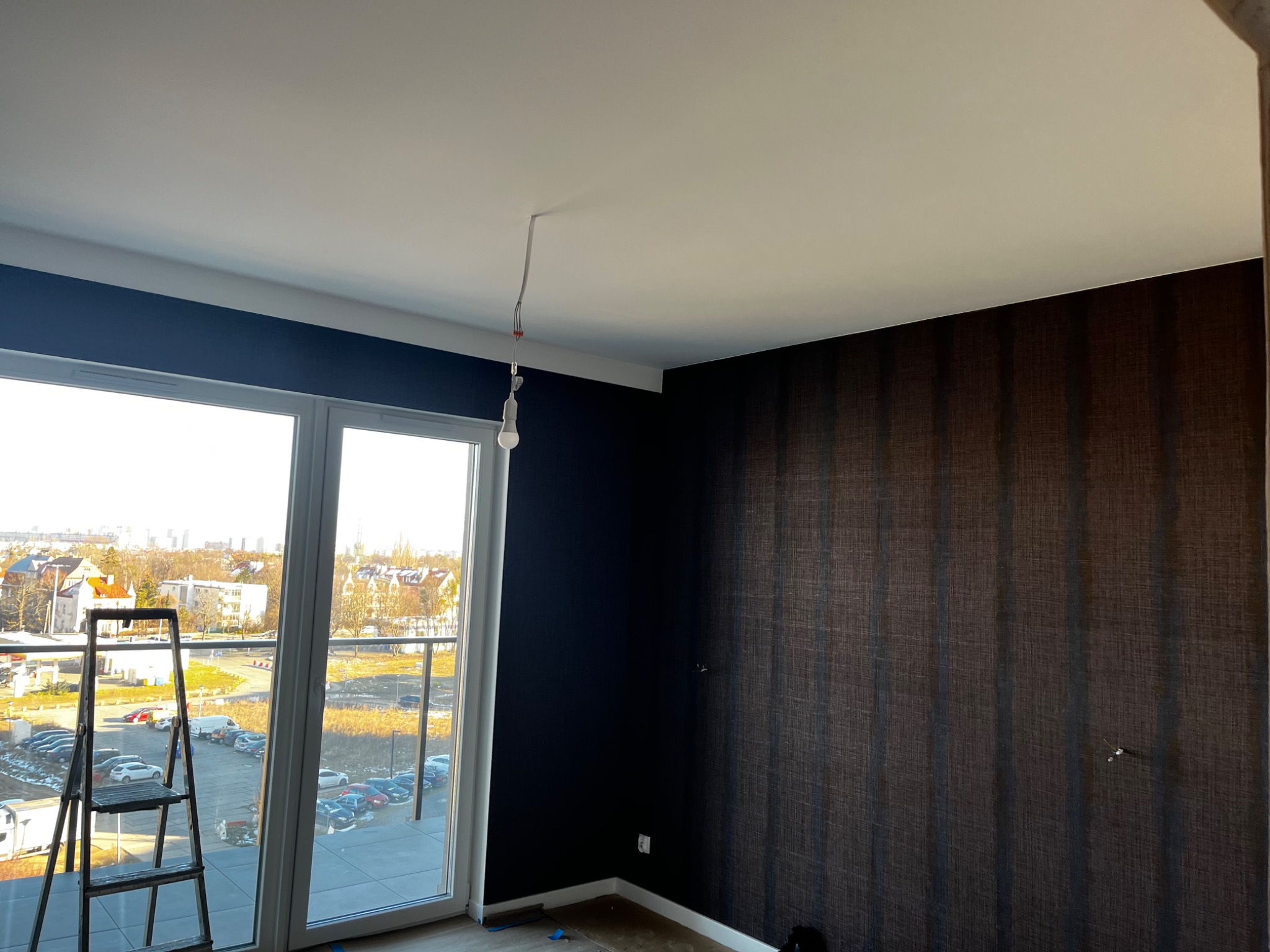 Pokój przed wykończeniem: ciemna ściana z tapetą w pionowe pasy, niebieska ściana, biały sufit z wiszącą żarówką, drabina przy oknie balkonowym z widokiem na miasto.