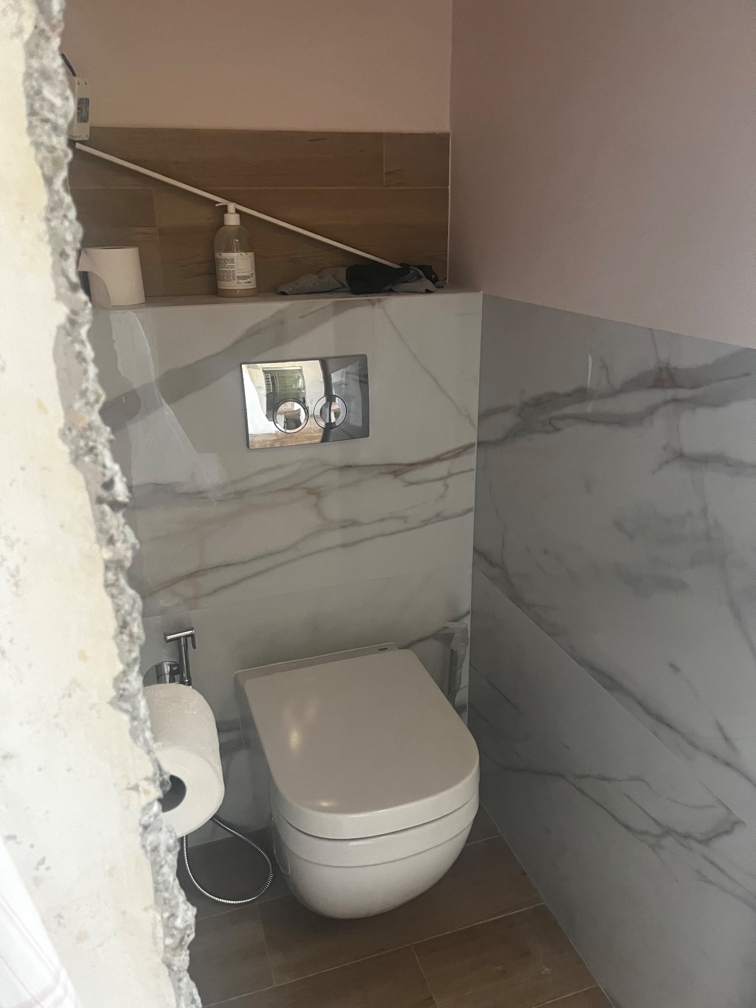 Podwieszana toaleta z marmurowymi płytkami w trakcie remontu łazienki, widoczne surowe krawędzie ściany i fragment instalacji.