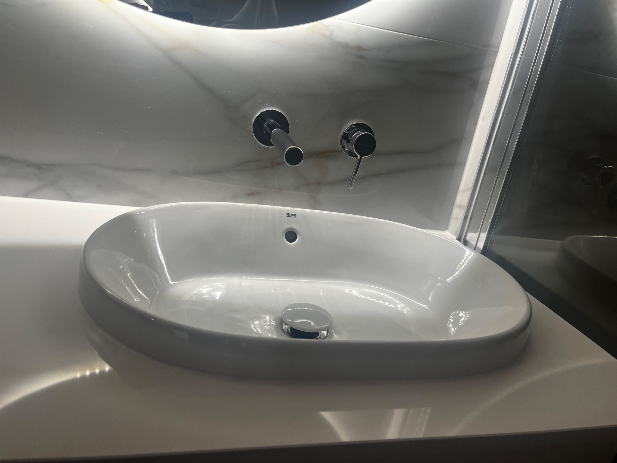 Nowoczesna, owalna umywalka Roca zamontowana na białym blacie w łazience z marmurową ścianą i chromowaną armaturą.