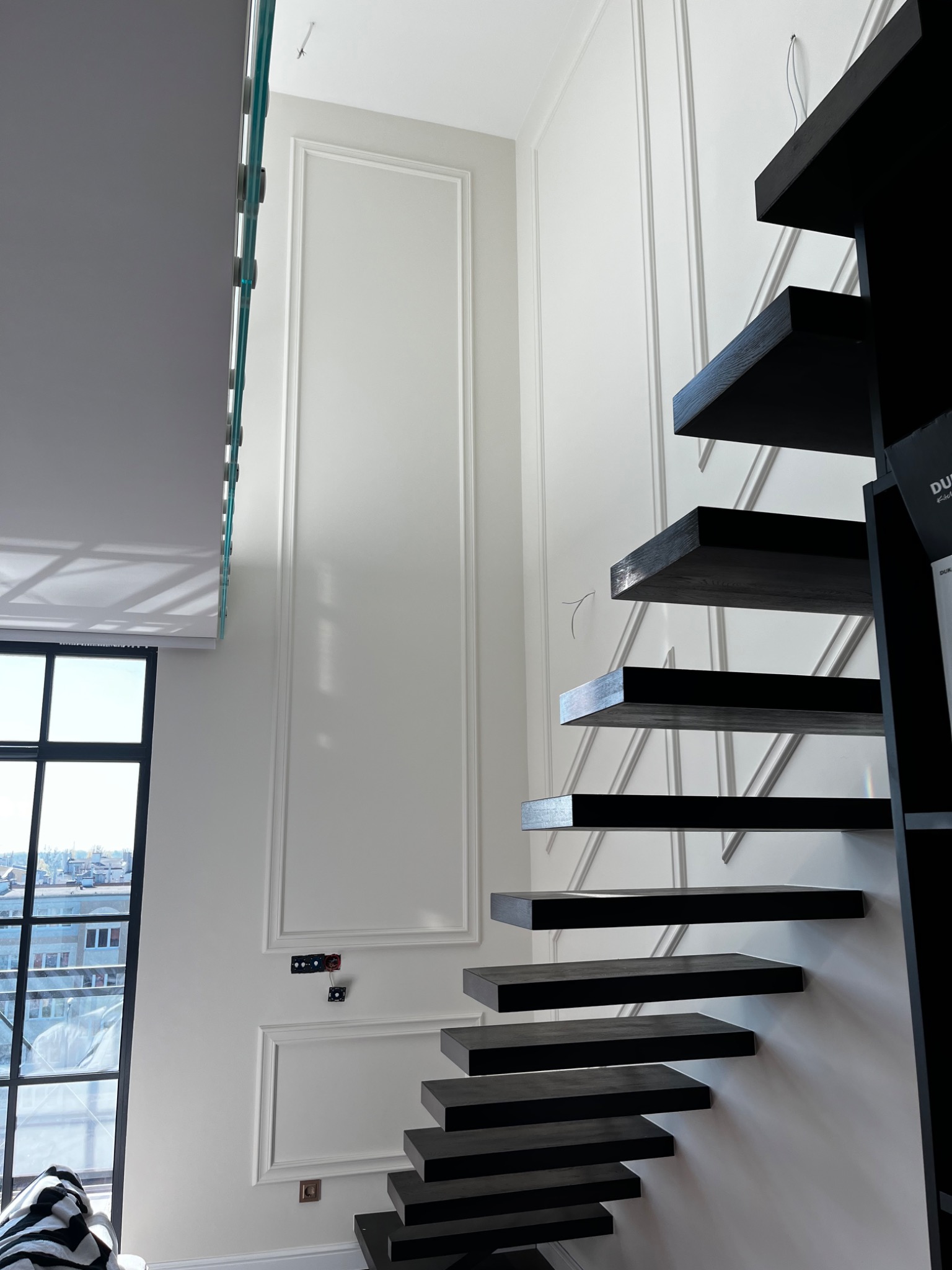 Nowoczesne, czarne, drewniane schody wspornikowe przy białej ścianie z dekoracyjnymi listwami i widocznymi puszkami elektrycznymi, z oknem w tle.