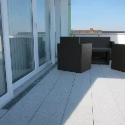 Nowoczesny taras z jasnoszarych płyt, wyposażony w plecione meble ogrodowe i odpływ liniowy przy oknie balkonowym.