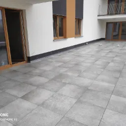 Nowoczesny taras wyłożony szarą, kwadratową płytką z widocznymi drzwiami balkonowymi i elementami elewacji budynku.