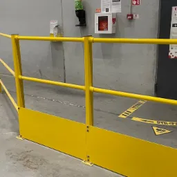Żółta, stalowa barierka ochronna z dolną osłoną, zamontowana w hali przemysłowej, z widocznymi oznaczeniami ostrzegawczymi na podłodze i plakatem informacyjnym na ścianie.