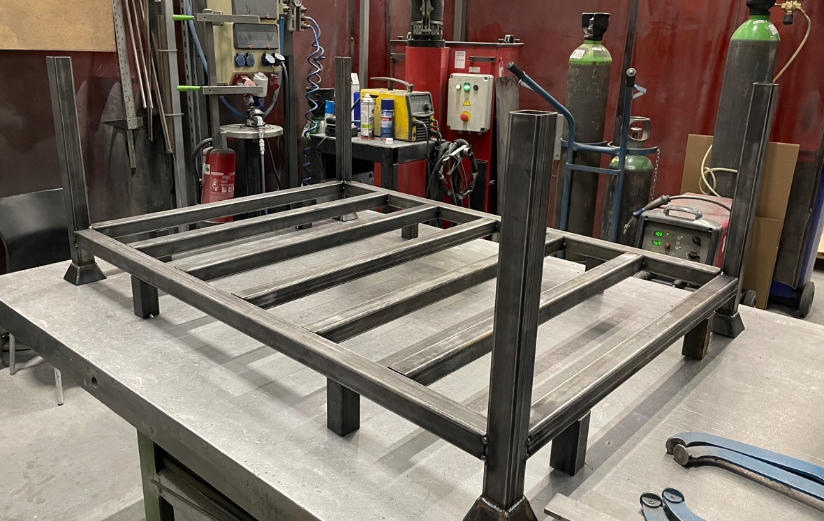 Stelaż z profili stalowych spawany na stole warsztatowym, widoczne spawy, narzędzia i butle z gazem w tle.