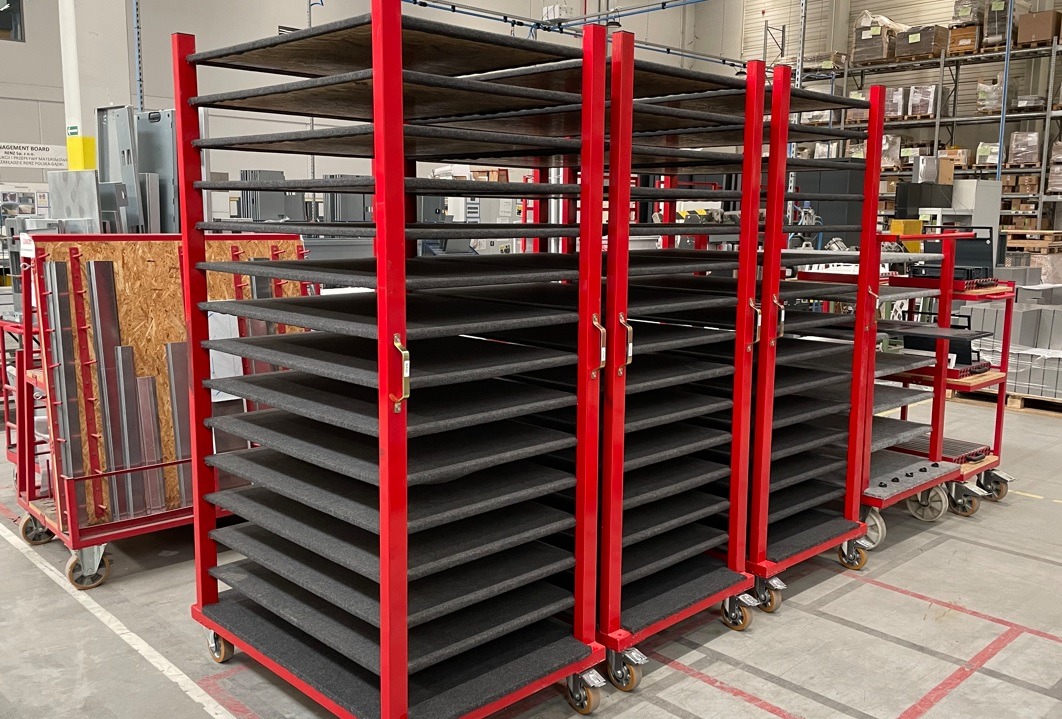 Metalowe wózki z wieloma poziomami, pomalowane na czerwono, z szarymi, wyściełanymi półkami, stojące w hali produkcyjnej.