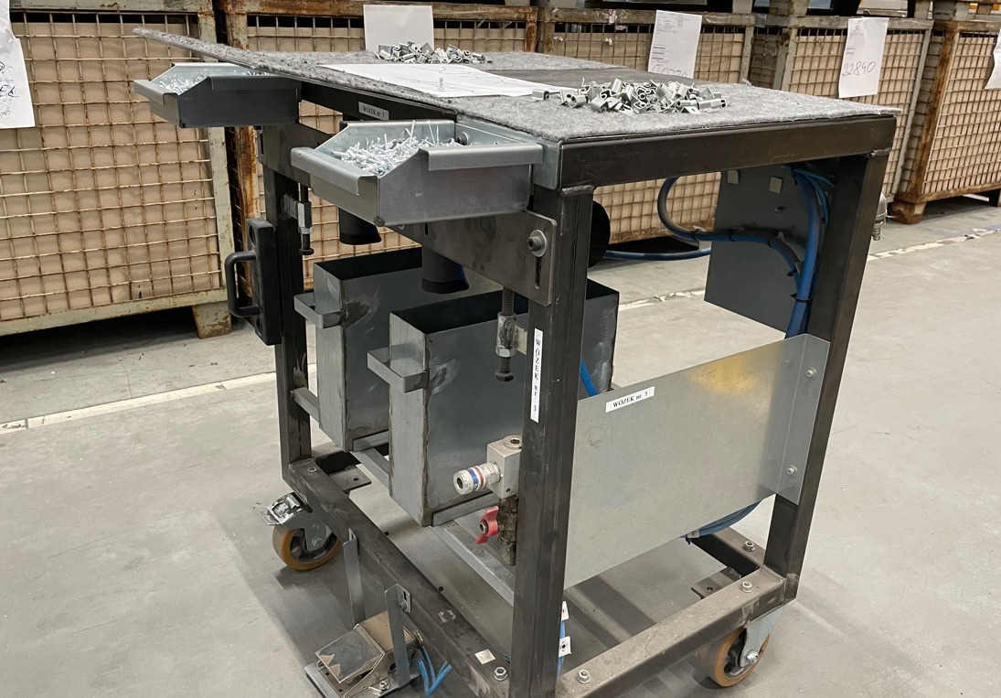 Wózek warsztatowy z metalową konstrukcją, wyposażony w pojemniki na drobne elementy montażowe, widoczne śruby, klamry i inne akcesoria, na górze wyłożony szarą wykładziną, całość na kółkach.