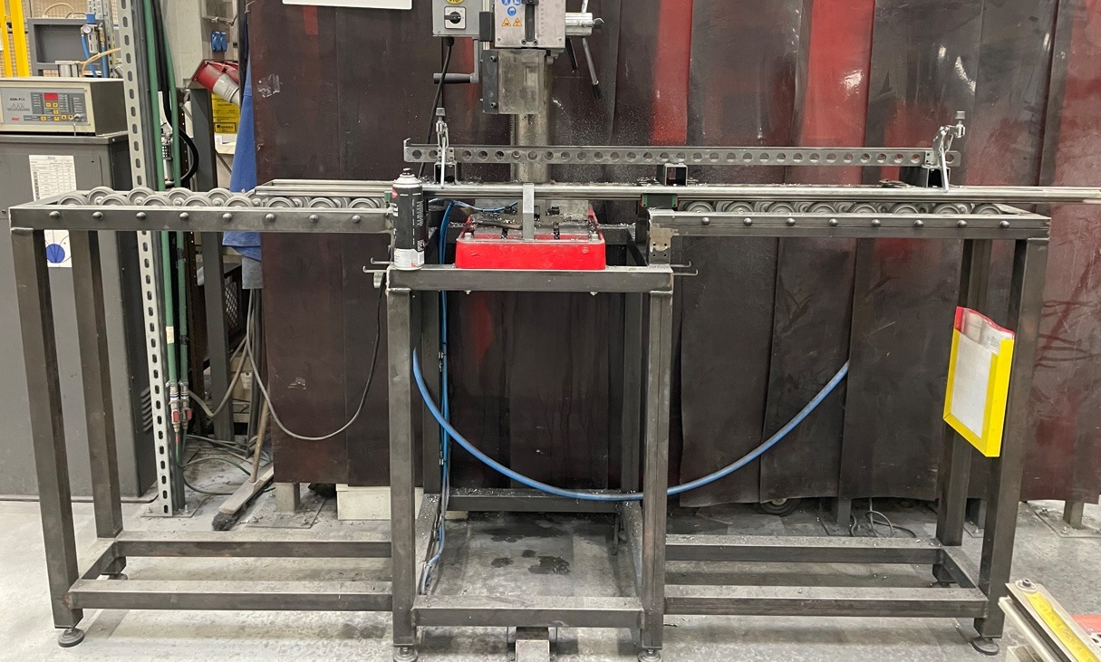 Stół roboczy z metalową konstrukcją i rolkami transportowymi, z widocznym urządzeniem do obróbki metalu w tle przesłoniętym kurtyną spawalniczą.