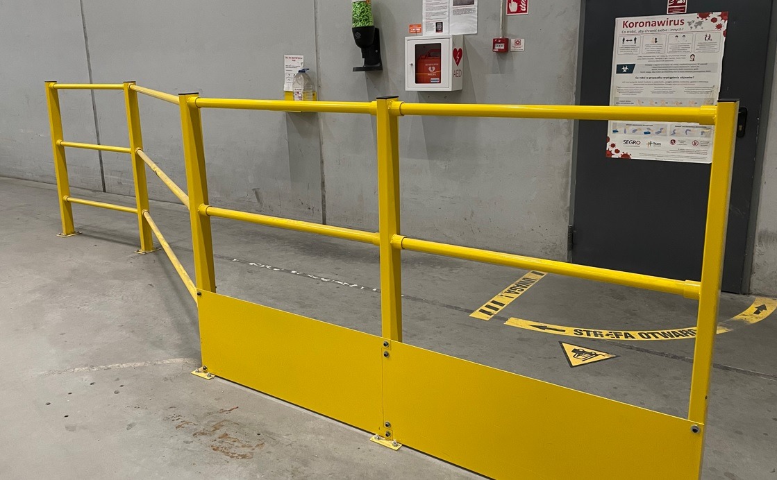 Żółta, stalowa barierka ochronna z dolną osłoną, zamontowana w hali przemysłowej, z widocznymi oznaczeniami ostrzegawczymi na podłodze i plakatem informacyjnym na ścianie.