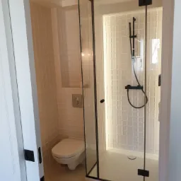 Nowoczesna łazienka z kabiną prysznicową typu walk-in z czarnymi profilami, wiszącą toaletą i białymi płytkami imitującymi cegiełki.