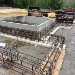 Wykop pod budynek z zalewanymi betonem fundamentami, widoczna zbrojona konstrukcja i woda w wykopie, na dalszym planie składowane materiały budowlane.