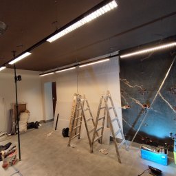 ELKOR Figurski Sp. z O. O. - Montaż liniowego oświetlenia LED w pomieszczeniu z marmurową ścianą, widoczne drabiny i narzędzia.