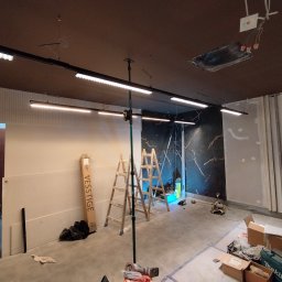 ELKOR Figurski Sp. z O. O. - Wnętrze w trakcie montażu oświetlenia LED na szynoprzewodach, widoczne drabiny, narzędzia, niedokończone ściany i sufit z odsłoniętymi przewodami.