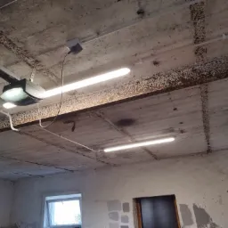 Wnętrze garażu z surowym betonowym stropem, widoczne dwie świetlówki, automat do bramy garażowej, instalacja elektryczna natynkowa i fragment ściany z oknem.