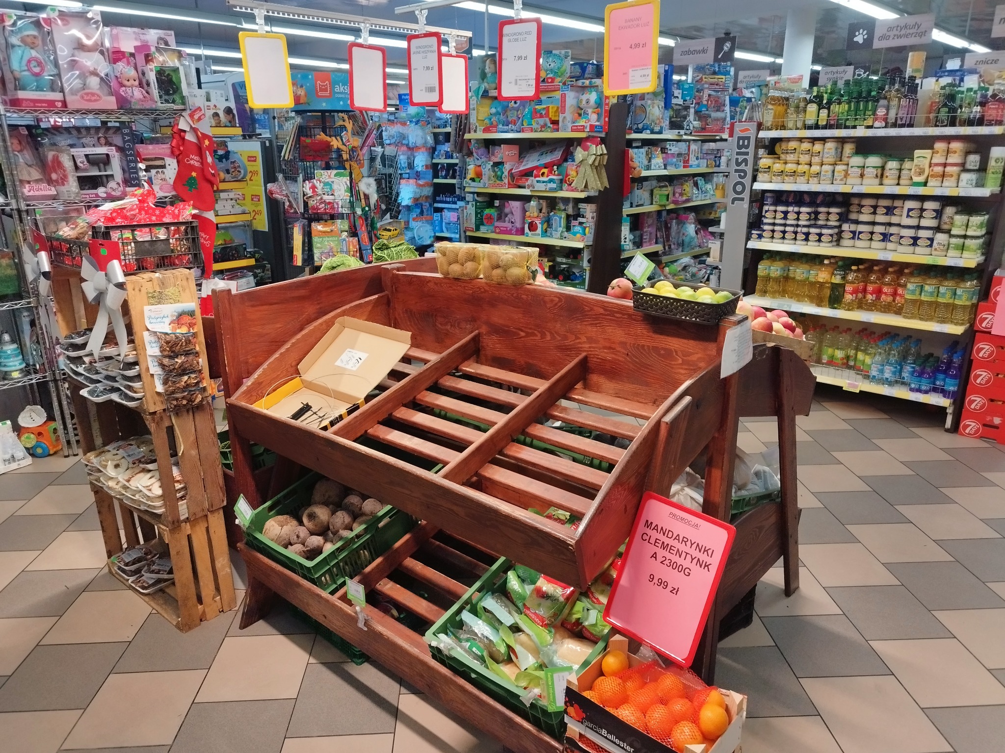 Półki sklepowe z drewnianą konstrukcją, zawierające owoce i warzywa, w tle regały z zabawkami i artykułami spożywczymi, widoczne ceny promocyjne.