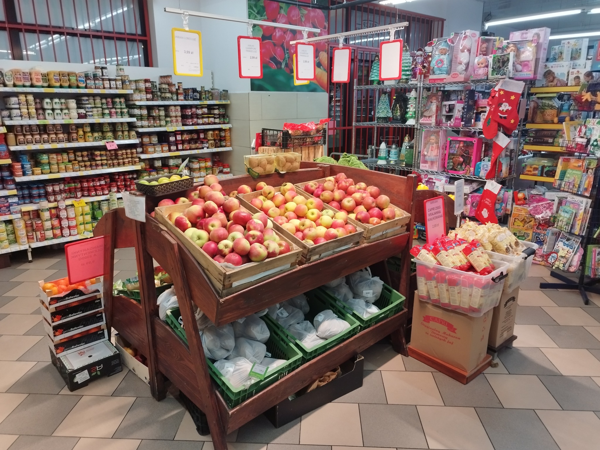 Stoisko z jabłkami, mandarynkami i artykułami świątecznymi w supermarkecie, widoczne ceny i opakowania produktów.